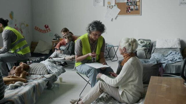 Full shot of volunteers in yellow vests assisting people displaced due to war or disaster in refugee shelter. Caucasian woman with clipboard sitting on chair, questioning senior lady about her needs