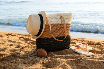 Stylish beach bag, coconut, hat and flip-flops on sand near sea