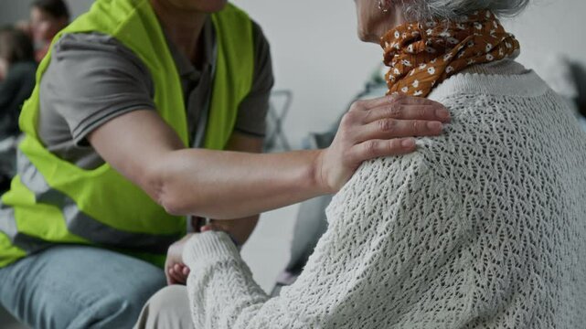 Medium shot of unrecognizable female volunteer in yellow vest consoling elderly lady in refugee shelter, displaced due to war or natural disaster, holding hand, stroking, patting on shoulder