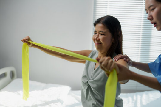 Asian senior woman doing physiotherapy exercises with resistance band assisted by nurse in hospital room
