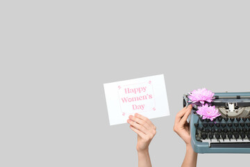Obraz premium Hands holding vintage typewriter, flowers and festive postcard with text HAPPY WOMEN'S DAY on grey background
