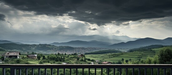 Fototapeta premium Dramatic cloudy landscape over rolling hills with distant mountains and a serene village in the foreground framed by a wooden railing.