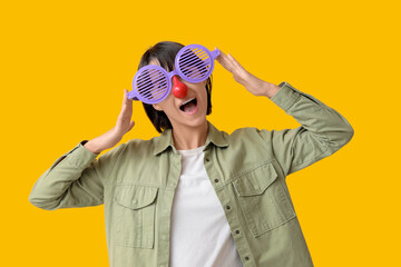Beautiful young shocked woman in funny disguise on yellow background. April Fools Day celebration