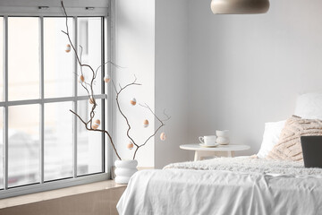 Vase with tree branches and Easter eggs on windowsill in light bedroom interior