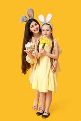 Happy smiling young woman with her daughter in bunny ears holding Easter cake and flowers on yellow background