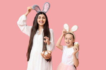 Happy smiling young woman with her daughter in bunny ears holding Easter cake and basket with eggs on pink background