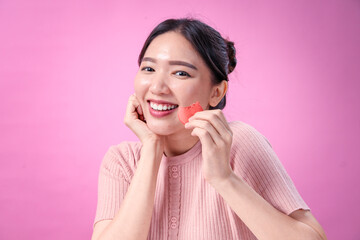 Fototapeta premium A young Asian woman with dark hair in a bun, wearing a ribbed pink short-sleeve top, smiles joyfully while holding a bitten red macaron, expressing happiness against a soft pink background.