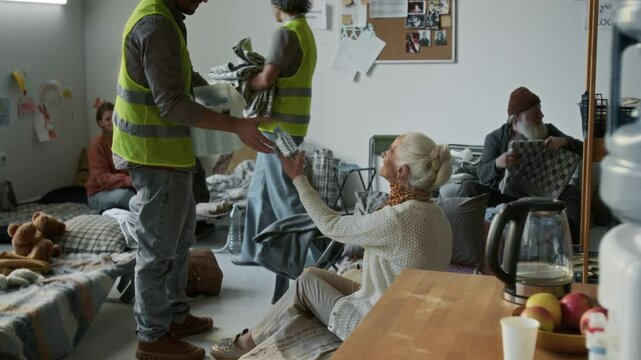 Full pan shot of volunteers in yellow vests distributing bottles of drinking water and blankets to mother, child, senior man and elderly woman, affected by natural disaster, staying in refugee shelter