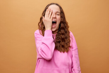 A woman with curly hair in a pink hoodie covers her face with one hand while joyfully laughing. The...