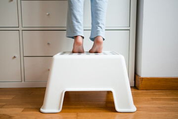 Caucasian child standing on white step stool reaching high cabinet