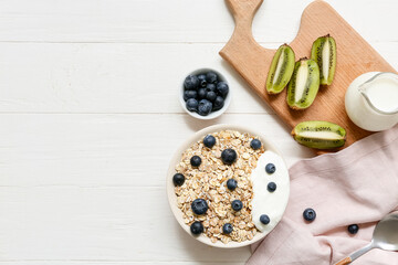 Napkin, wooden board, kiwi, bowl with raw oatmeal and berries on white wooden background