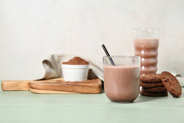 Glasses of sweet chocolate milk with tasty cookies and bowl of cocoa powder on green wooden table