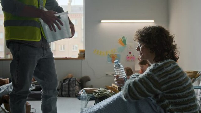 Medium full shot of anonymous male volunteer in yellow vest giving bottle of water to depressed woman, displaced by natural disaster or war, sitting on mattress in refuge, woman thanking him