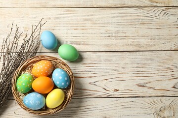 Beautiful decorated Easter eggs, wicker bowl and willow branches on light wooden table, flat lay. Space for text
