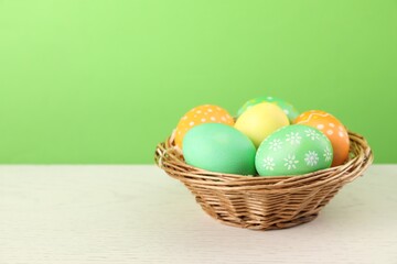 Many decorated Easter eggs in wicker basket on wooden table against light green background, space for text