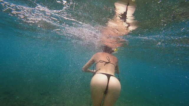 Woman snorkeler swimming underwater in crystal-clear turquoise water. Person wearing bright yellow diving mask and snorkel with black swimsuit. Camera fast going to her simulates a shark attack