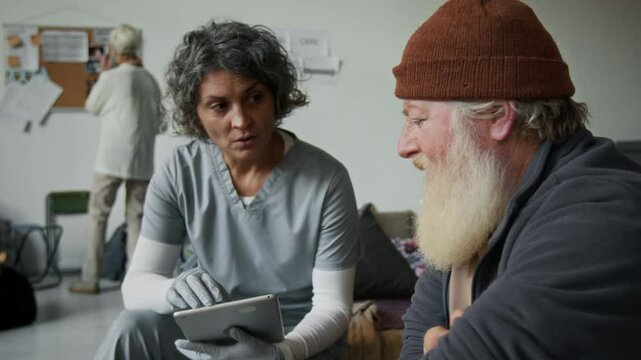 Medium shot of middle-aged Caucasian female doctor or nurse in medical uniform sitting in refugee shelter, filling checklist on tablet, talking to senior male disaster survivor about health issues
