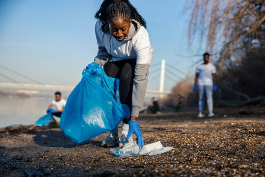 Interracial ecologist collecting plastic waste on riverbank with her team in a blurry background on earths day.