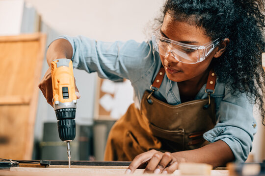 Carpenter woman using a power drill to secure a piece of wood in a workshop, focusing on precision in furniture making. Surrounded by tools and wood pieces, highlighting skill and craftsmanship
