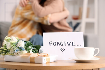 Paper with word FORGIVE, coffee cup and gifts on table against hugging couple at home, closeup