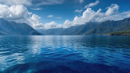 Fototapeta premium Tranquil deep blue ocean reflecting mountains under a clear sky with fluffy clouds creating a serene and picturesque coastal landscape.