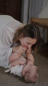 A little infant baby girl lies on the floor on a blanket, her mother hangs over her, takes her feet in hands and kisses them tenderly with love. Maternity leave. Mothers Day.