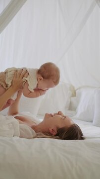 The young woman mother lies on the white bed and lifts her little daughter on the hands. They play and laugh. Maternity leave lifestyle. The woman tenderly kisses her infant daughter. Mothers day.