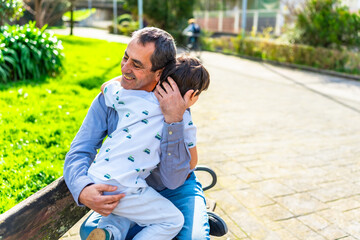 Fototapeta premium Grandfather hugging grandson while sitting on bench in park