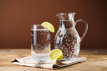 Refreshing soda water with ice cubes in glass, jug and lemon slices on wooden table against brown background