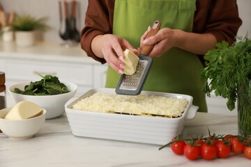 Woman grating cheese onto spinach lasagna at marble table indoors, closeup
