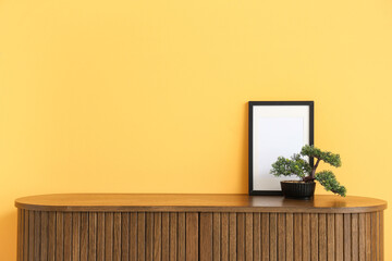 Blank frame with bonsai tree on commode near yellow wall