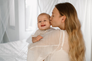 A young woman holds her little daughter on the arms and kisses her tenderly. Adorable baby smiles happily. Rocks the baby to sleep. Mothers Day. Love and affection.