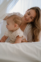 Mother and her baby daughter on a bed on a white blanket. The infant baby is crawling on the bed, and woman is talking to her and smiling. Everyday life of a mother on maternity leave. Mothers Day.