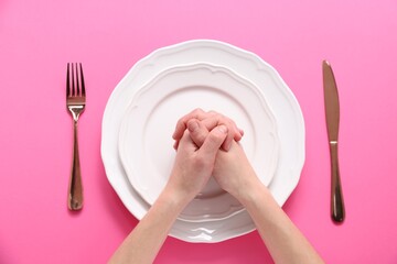 Woman holding hands together over plate and cutlery on pink background, top view