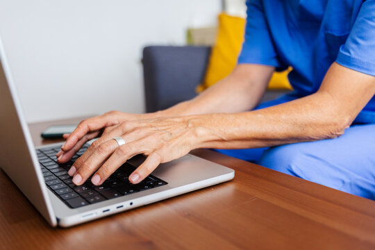 Close up view of unrecognizable senior doctor working on laptop sitting on sofa at home after workday in hospital. Technology and healthcare business concept.