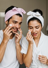 Image of young loving couple in bathrobes smiling and using cotton pad while standing in bathroom. Couple Skincare Routine