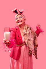 Elderly woman with joyful expression wearing pink outfit, holding glass of milk over pink background. Confident charm. Fashion pop art photography. Concept of playful elegance, celebration.