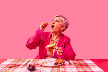 Elderly woman in pink suit eating croissant against pink studio background. Lively breakfast scene. Food pop art photography. Concept of fashion and food, leisure and enjoyment