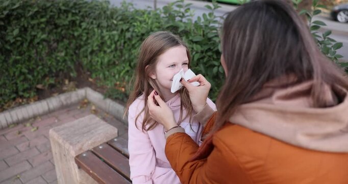 Caring mother uses tissue to wipe nose of daughter. Parent wipes runny nose of child with tissue during walk in autumn city park. Slow motion