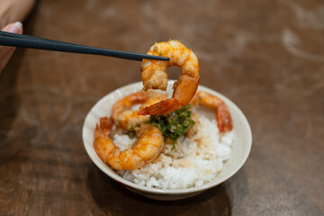 Chopsticks lifting a shrimp above a bowl of stir-fried shrimp and white rice.