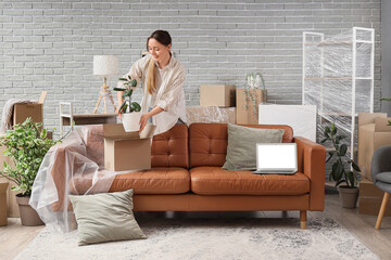 Young woman putting plant into moving box and blank laptop on sofa in room