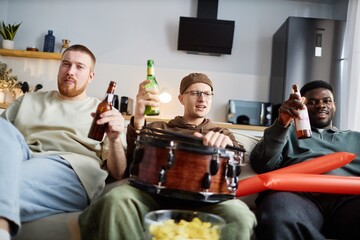 Group of cheerful sports fans with drinks and instruments sitting on couch while watching game on TV at home, copy space