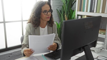 Middle-aged woman in an office reads documents while working at a computer with focus and calm demeanor in a corporate indoor environment.