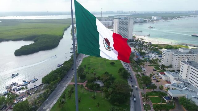 Mexican flag flying on a flagpole in the median of the Cancun Quintana Roo hotel zone, beautiful waves, sea and beach are seen in the background, as well as the Nichupt&eacute; lagoon on a beautiful day