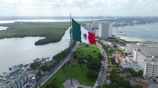 Mexican flag flying on a flagpole in the median of the Cancun Quintana Roo hotel zone, beautiful waves, sea and beach are seen in the background, as well as the Nichupt&eacute; lagoon on a beautiful day