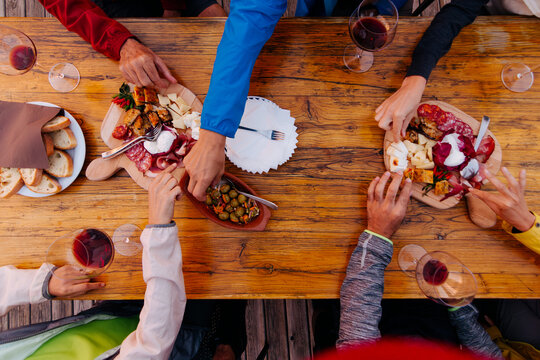 Friends sharing a rustic meal together outdoors - group of people enjoying a shared meal with wine, cheese, bread, and charcuterie on a wooden table