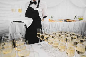 A waiter in formal attire holds a tray with champagne glasses, while a table in the background...