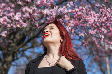 Red haired woman wearing stylish outfit near blossoming sakura in park. Fashionable spring look....
