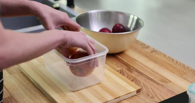 Woman hands take washed fruits from metal bowl at wooden countertop in kitchen. Housewife puts apples into transparent container for food slow motion