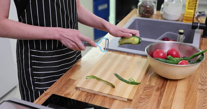 Housewife in apron uses vegetable peeler peeling cucumber standing at wooden countertop. Female prepares vegetable for salad in kitchen slow motion